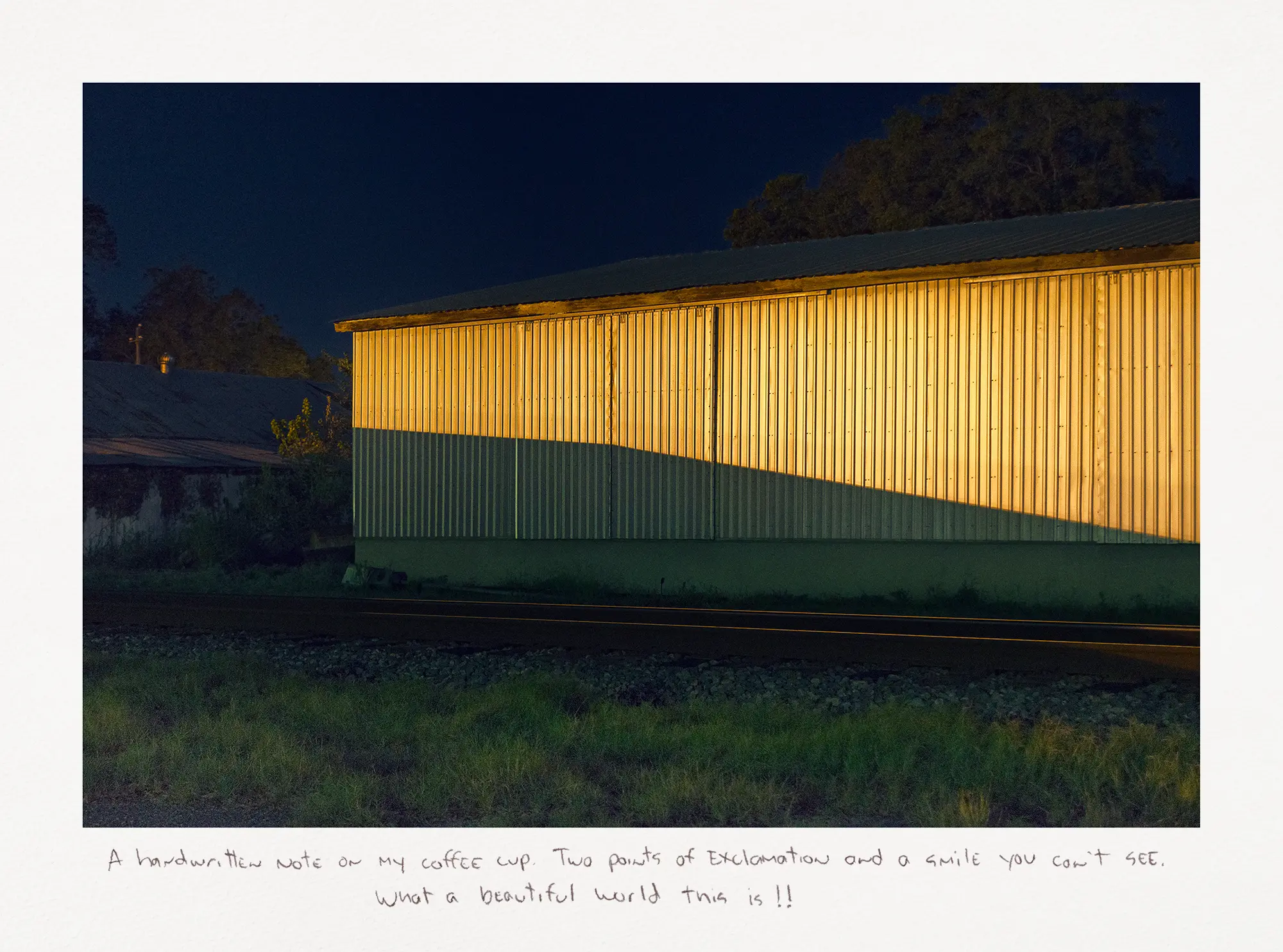 A photograph of a corrugated building beside railroad tracks illuminated by orange light.